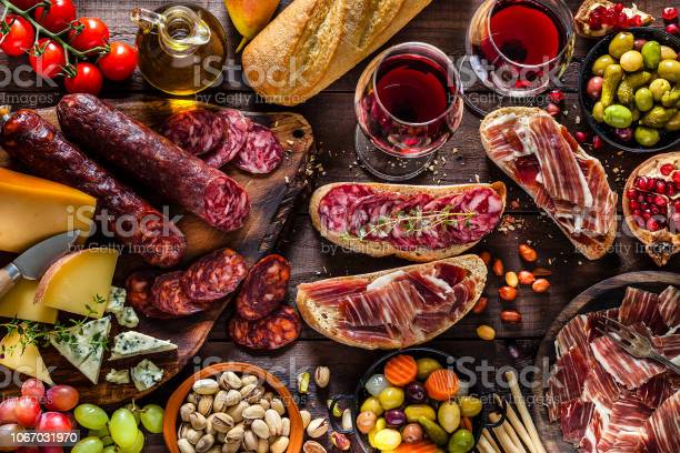Top view of a rustic wooden table filled with delicious cheese and cold meat selection. Some fruits like figs, pears and grapes, olives, pistachios, bread, breadsticks an olive oil bottle and two wineglasses complete the composition. DSRL studio photo taken with Canon EOS 5D Mk II and Canon EF 100mm f/2.8L Macro IS USM