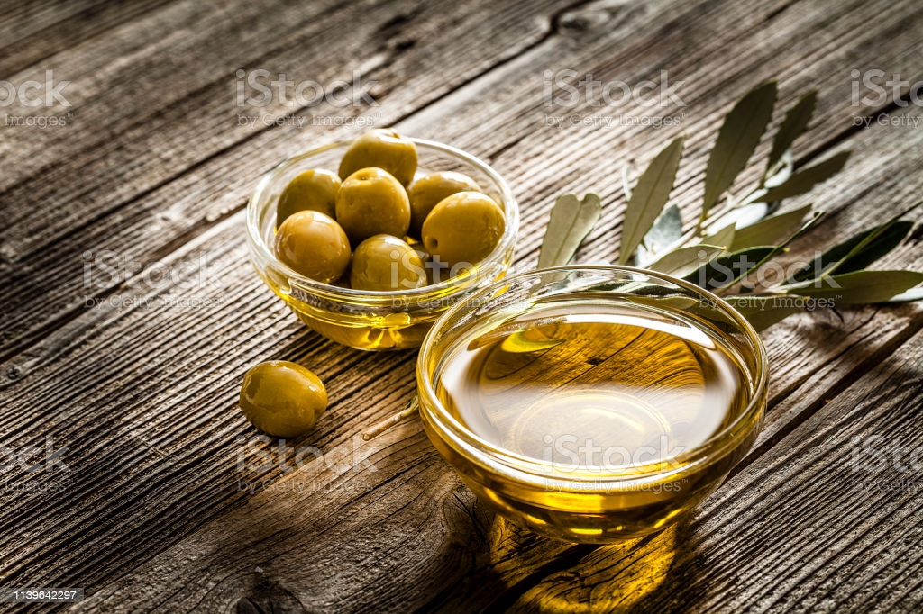 High angle view of a glass bowl filled with olive oil and green olives shot on rustic wooden table. Olive branches complete the composition. Predominant colors are brown and gold. Low key DSRL studio photo taken with Canon EOS 5D Mk II and Canon EF 100mm f/2.8L Macro IS USM.